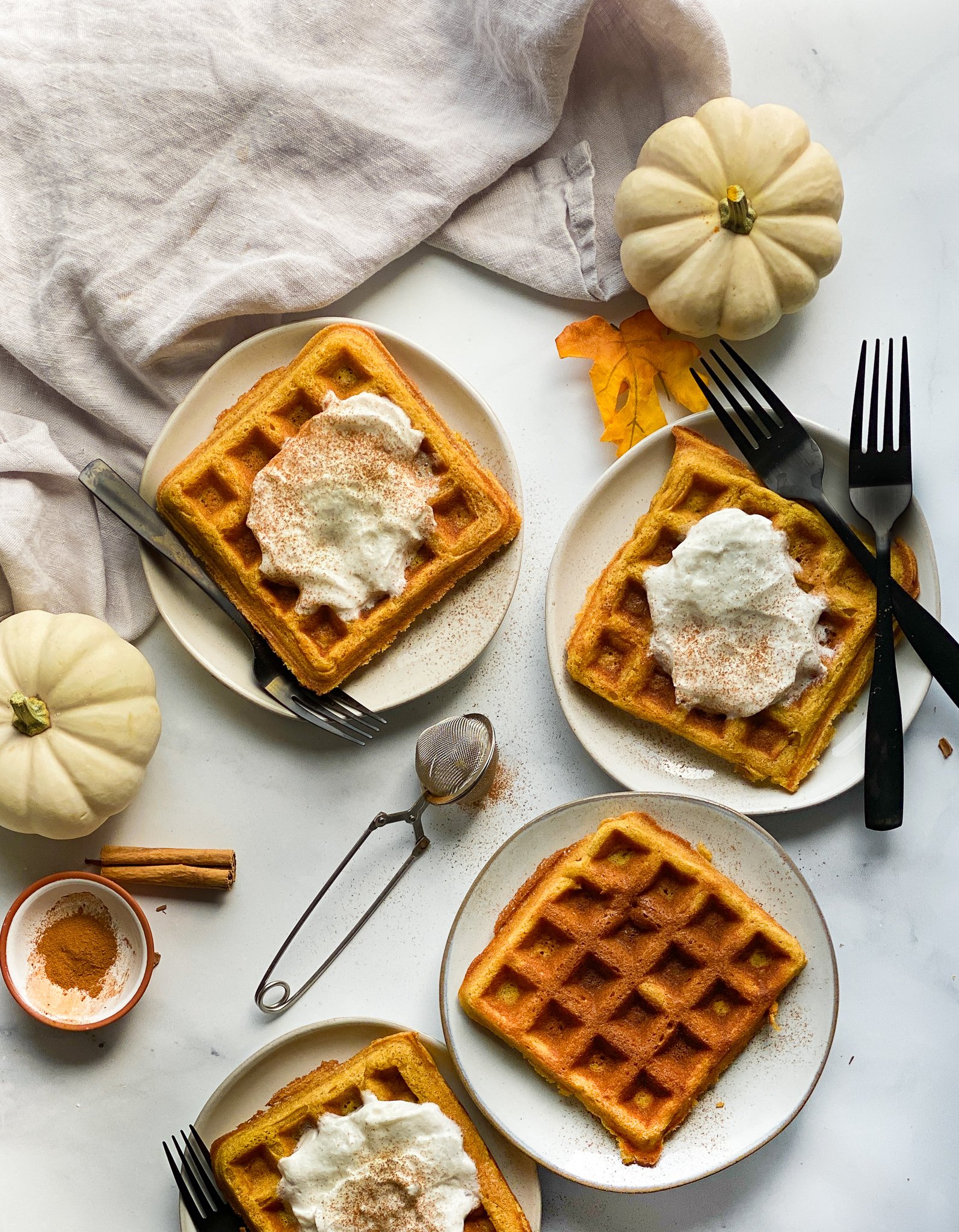 Grain-free pumpkin waffles sitting on a marble counter top on grey plates. There are four different plates and three of them have a dollop of fresh whipped cream and a dash of cinnamon on them.