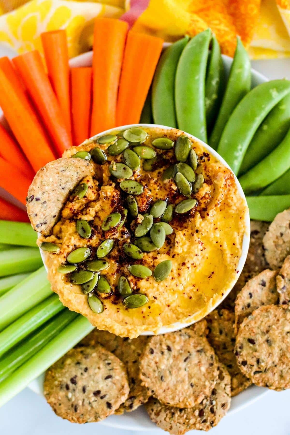 Bowl of pumpkin hummus garnished with pumpkin seeds and spices on a platter of celery, carrots, sugar snap peas, and crackers.