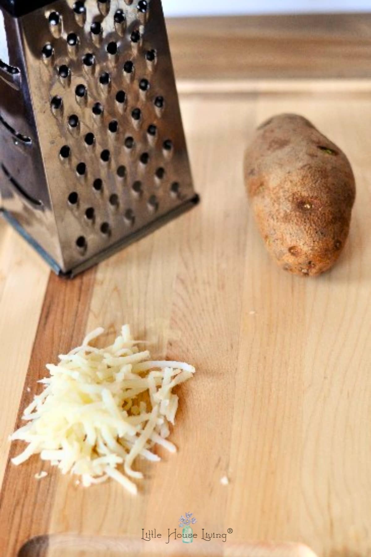 a box grater, whole potato and pile of shredded potato on a wooden cutting board